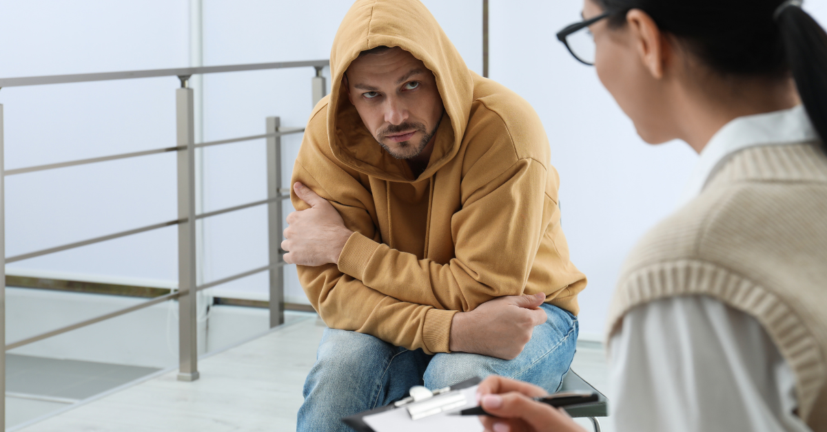 A man in a yellow hooded sweatshirt, talking to a woman with glasses while in opiate addiction treatment