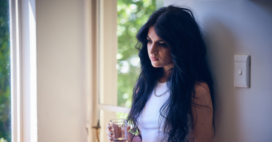 Woman with long dark hair leaning against a wall contemplating inpatient treatment vs outpatient treatment while holding a glass of water