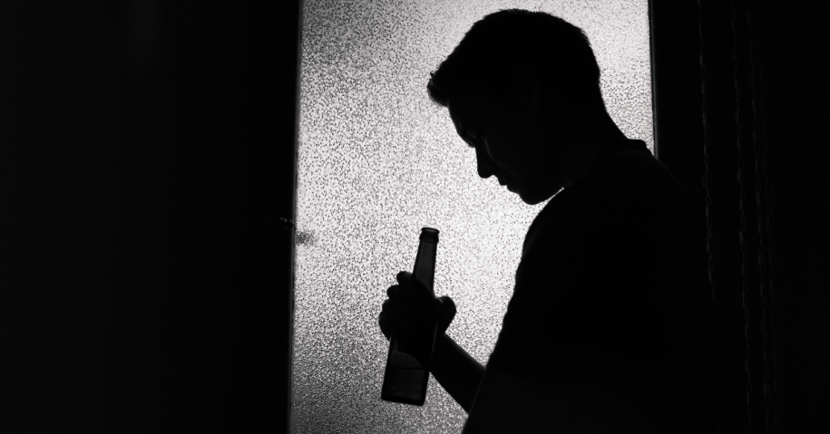 A man standing behind a door with a beer bottle in hand and his head tipped downward